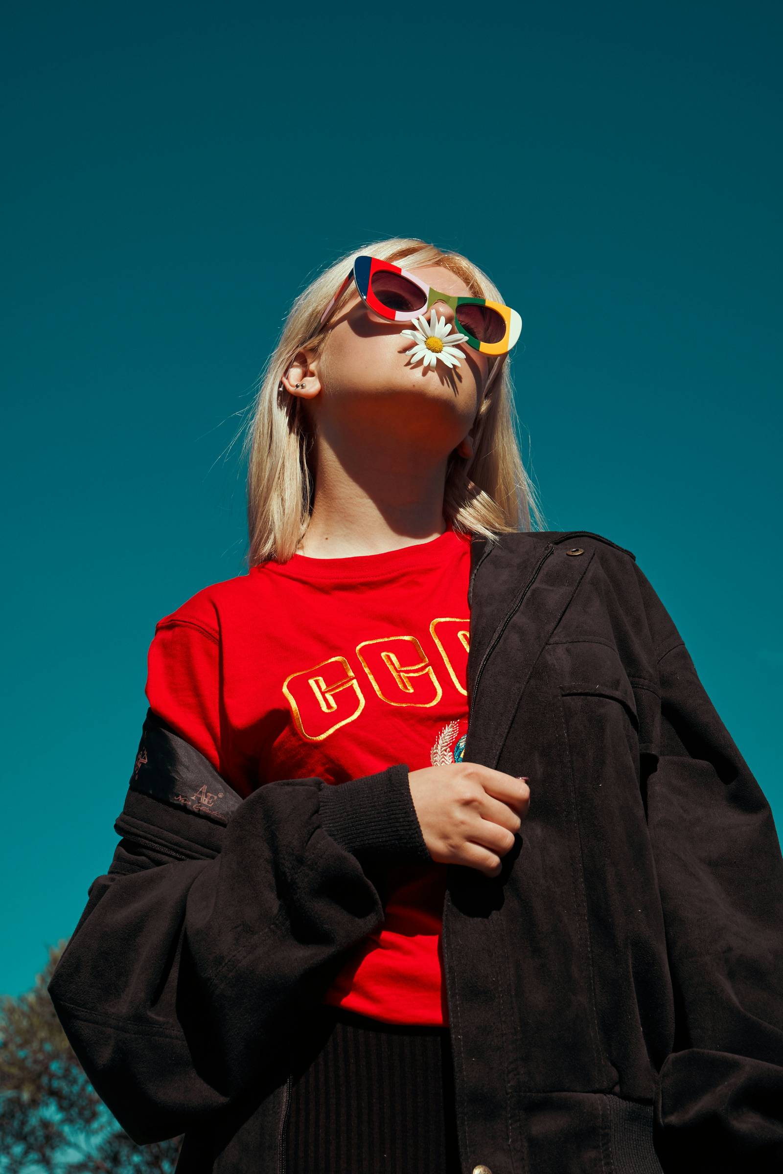 Sterling Tiger Valley Young woman in colorful sunglasses and red shirt holding a daisy in her mouth, standing under a clear blue sky.