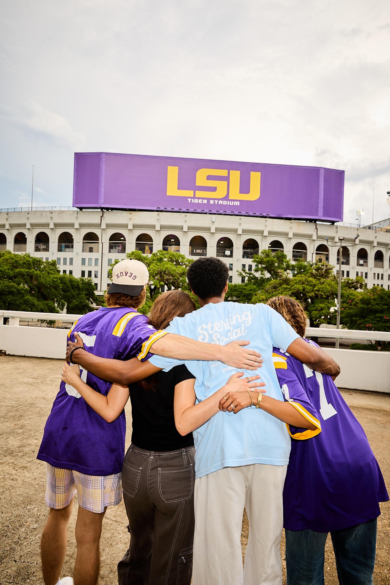 Sterling Tiger Valley Four people with arms around each other stand facing LSU Tiger Stadium, wearing casual and LSU-themed clothes.