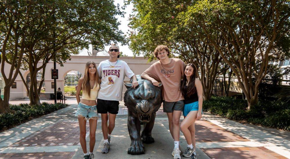 Sterling Tiger Valley Four people pose by a tiger statue on a tree-lined campus walkway under a sunny sky.
