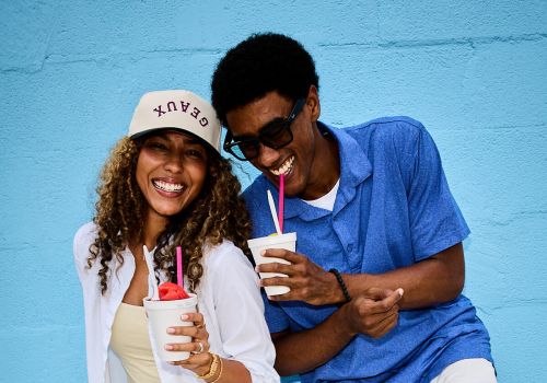 Sterling Tiger Valley Two people smiling and holding drinks with straws, standing against a bright blue wall, enjoying a casual moment together.