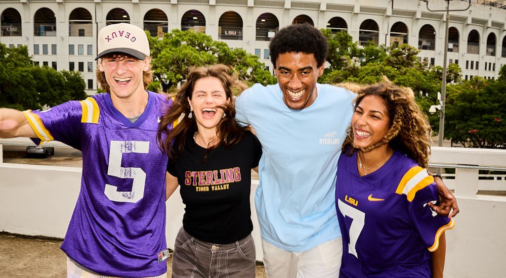 Sterling Tiger Valley Four smiling people pose in front of LSU Tiger Stadium, wearing LSU and sports-themed shirts and jerseys.