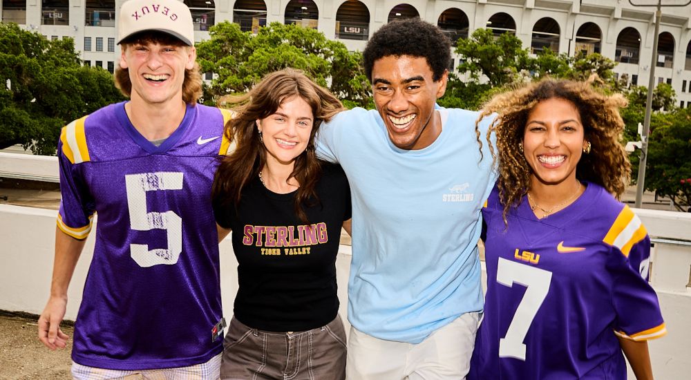 Sterling Tiger Valley Four young adults smile and pose in front of LSU Tiger Stadium, two wearing purple LSU football jerseys numbered 5 and 7.