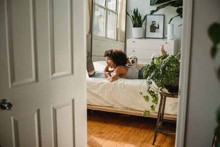 Sterling Tiger Valley A woman lies on her bed using a laptop with a dog beside her in a cozy, plant-filled room.