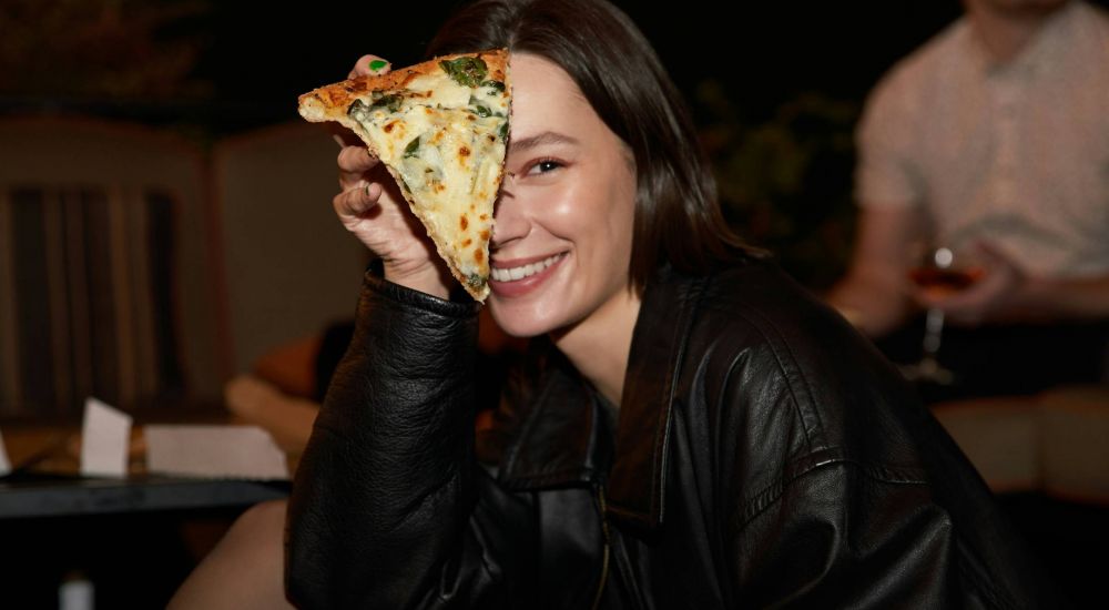 Sterling Tiger Valley Smiling woman holding a pizza slice over one eye while sitting outdoors at night.